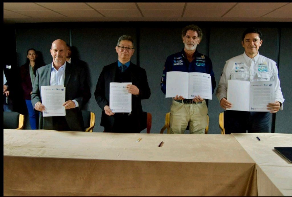 Four individuals holding signed documents during a signing ceremony, with additional people visible in the background.$# CAPTION