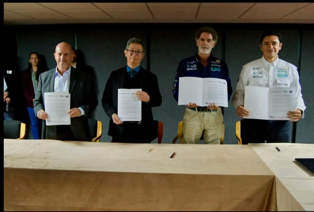 Four individuals holding signed documents during a signing ceremony, with additional people visible in the background.$# CAPTION