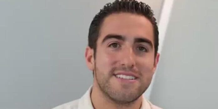 A young man with short dark hair and a light-colored shirt smiling in front of a neutral background.