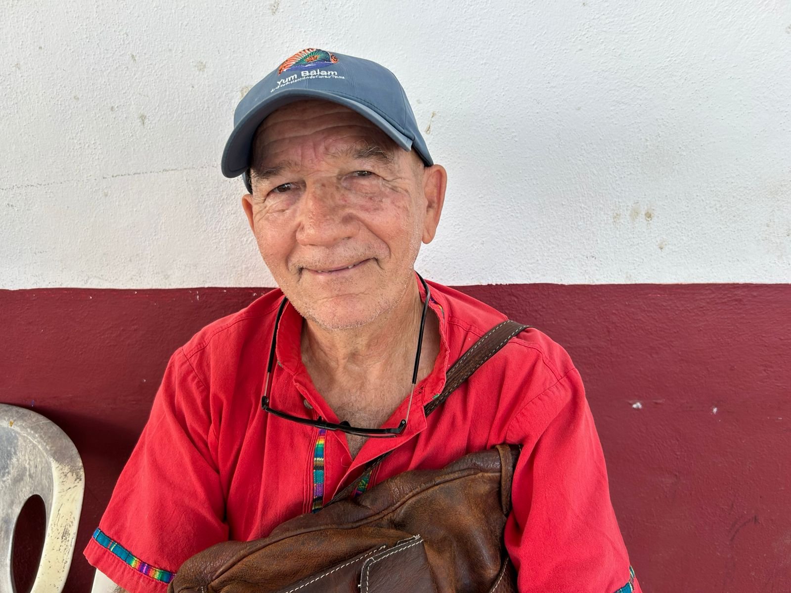 a smiling elderly man wearing a red shirt and a cap holding a leather bag while sitting against a wall with red and white colors