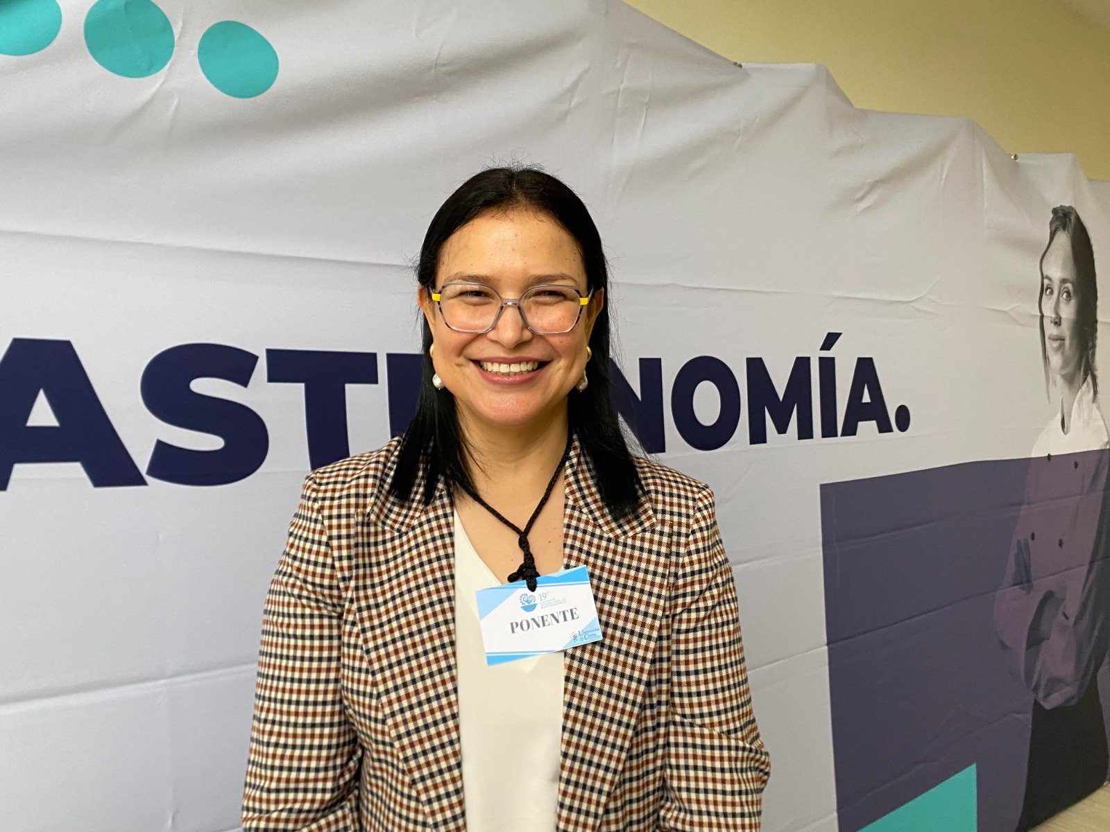 A woman with glasses in a checkered blazer stands in front of a backdrop that reads "ECONOMÍA" with a smiling expression. She has a name tag that says "PONENTE."