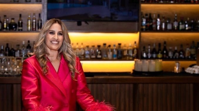 A woman in a vibrant pink suit with feather accents stands confidently at a bar, smiling. Behind her is a shelf stacked with various bottles of alcohol, illuminated by warm lighting.