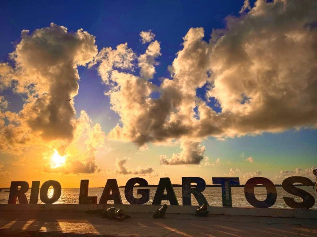 Colorful letters spelling 'Rio Lagartos' with a sunset and clouds in the background