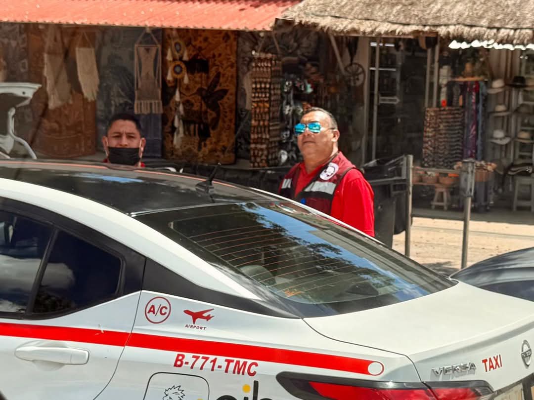 Two men at a taxi stand near a market, one wearing a black mask and the other wearing sunglasses, with various items visible in the background.