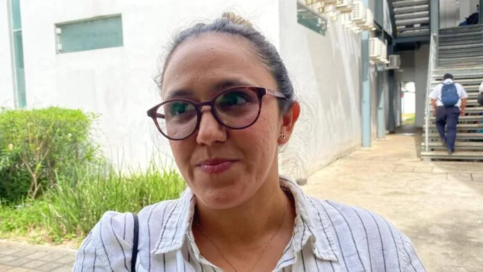 A woman with glasses, wearing a striped shirt, smiling softly while standing outside a building surrounded by greenery.$