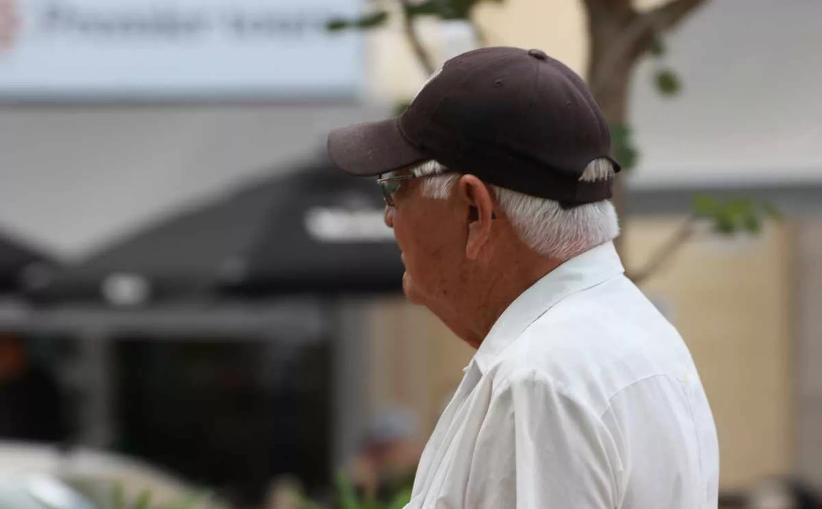 An elderly man wearing a cap and glasses is standing outdoors, looking away from the camera. His white hair is visible beneath the cap, and he is dressed in a white shirt. Soft focus background with trees and blurred restaurant umbrellas.$