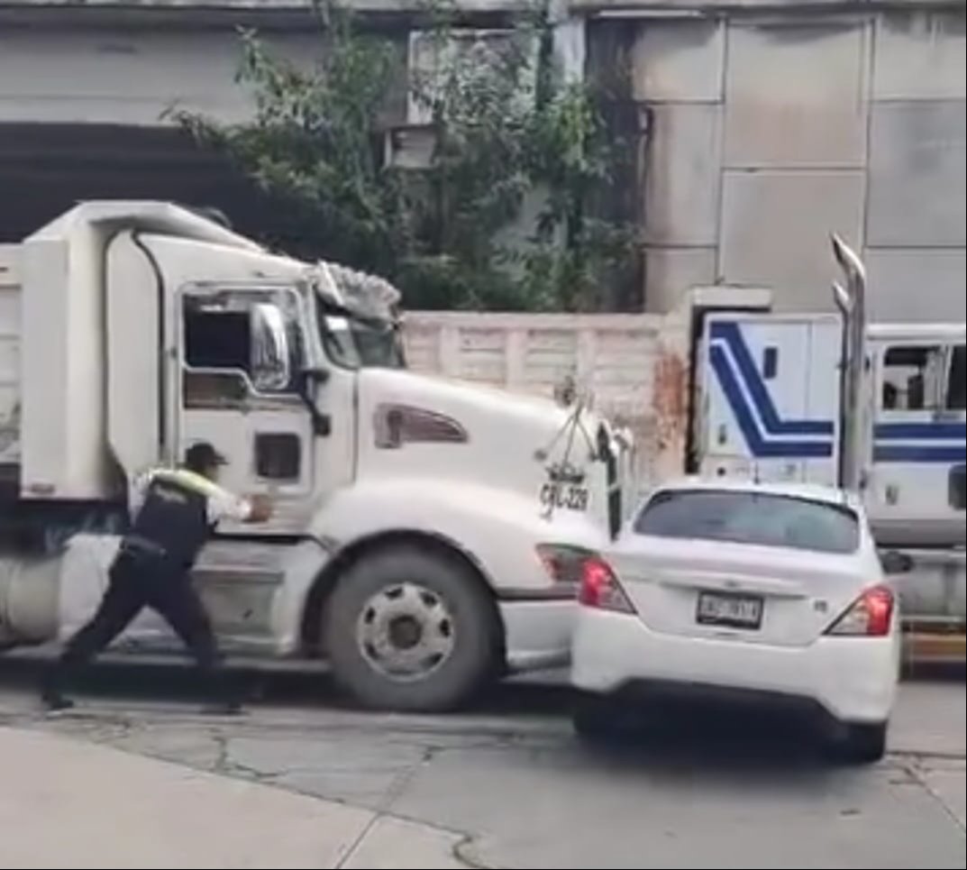 A truck driver attempts to maneuver their large vehicle while a smaller car is positioned closely behind it in a busy street setting. The driver appears to be pushing against the truck.