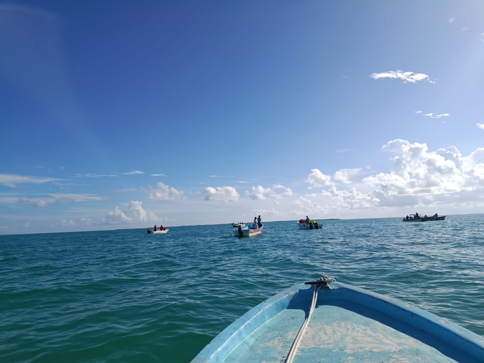 View from the bow of a blue boat looking out at several fishing boats on a calm ocean under a blue sky with scattered clouds