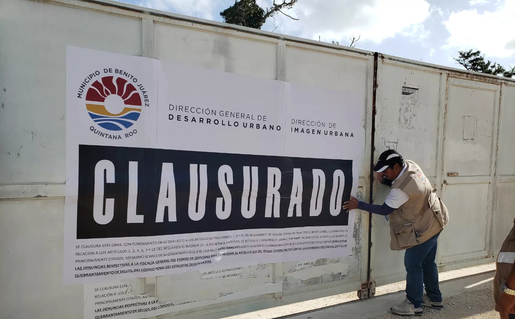A man affixes a large "CLAU- SURADO" (CLOSED) sign on a gate, indicating an urban development has been shut down in Benito Juárez, Quintana Roo, Mexico. The sign includes government logos and regulations.$# CAPTION