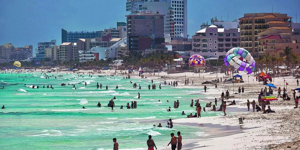 A lively beach with people enjoying the water and colorful parasails in the background, framed by city buildings and palm trees. The water is a striking turquoise color.