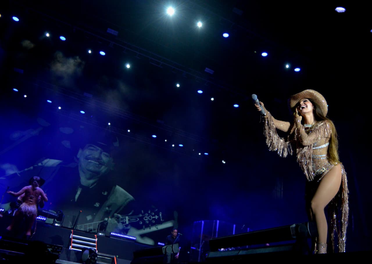 A singer in a glittering outfit performs on stage with dramatic lighting, while a black-and-white image of a man with a guitar appears in the background.