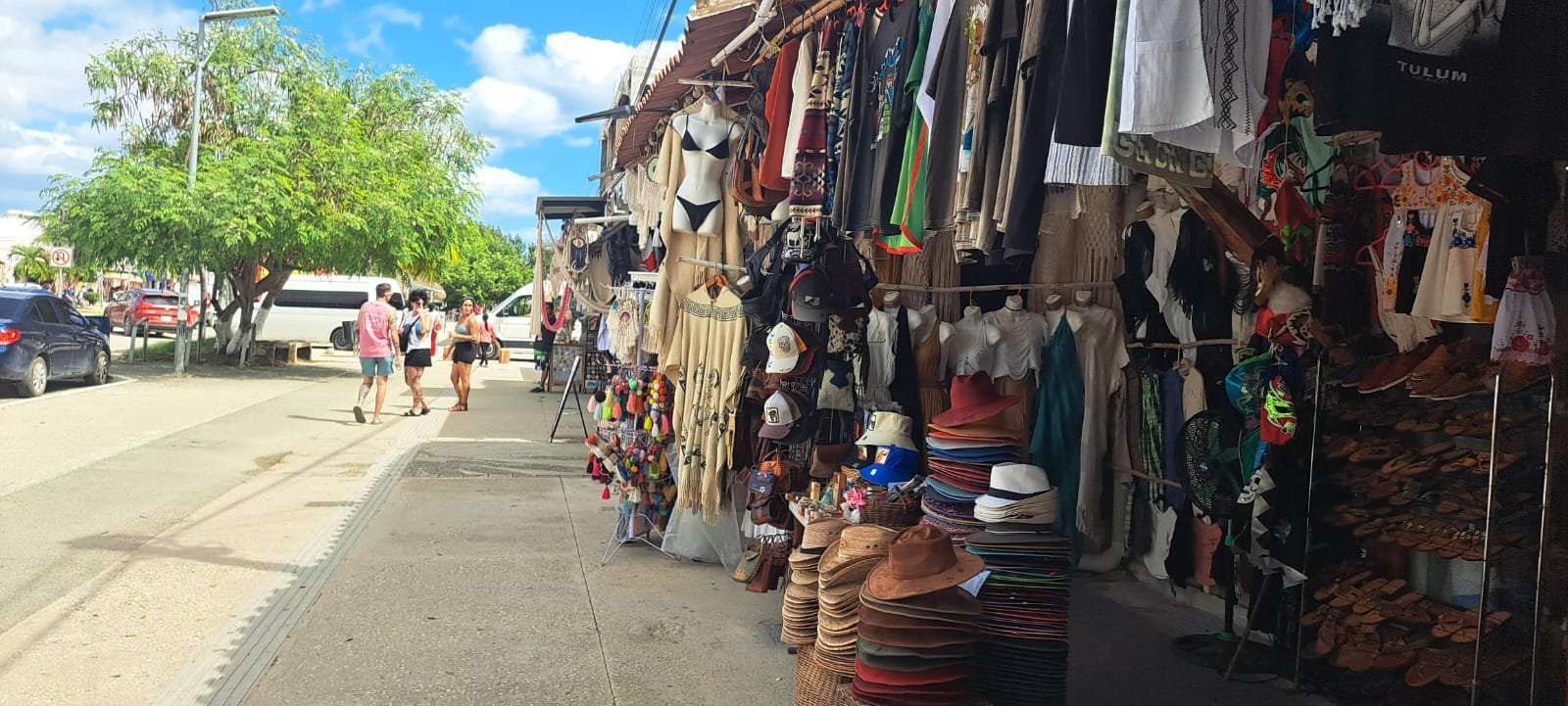 A bustling market street in Tulum filled with colorful clothing and accessories for sale, with shoppers walking by. A bright blue sky adds to the lively atmosphere.$# CAPTION