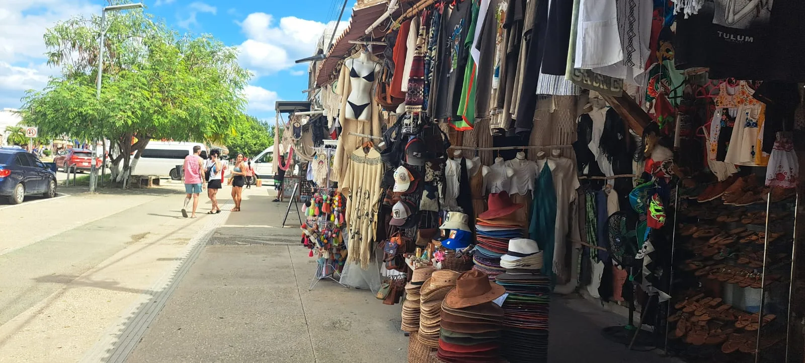 A bustling market street in Tulum filled with colorful clothing and accessories for sale, with shoppers walking by. A bright blue sky adds to the lively atmosphere.$# CAPTION