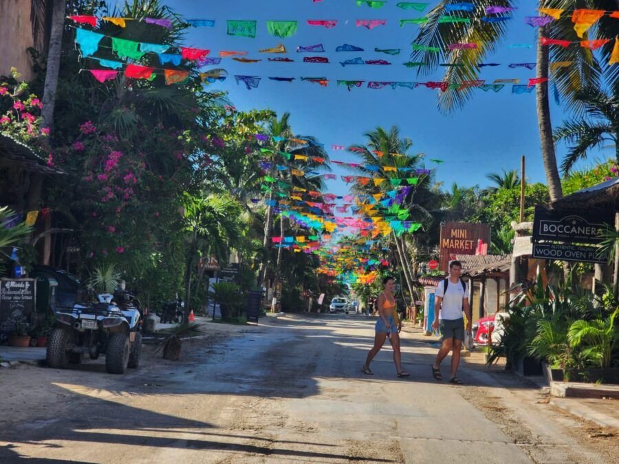 Colorful papel picado decorations hanging over a street lined with palm trees and shops, with two people walking.