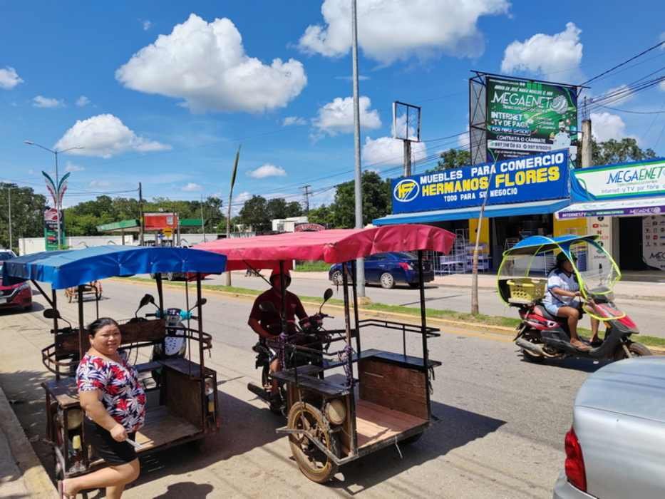 A lively street view showcasing colorful tuk-tuks and a woman standing on the sidewalk near a furniture store in sunny weather.$#$ CAPTION