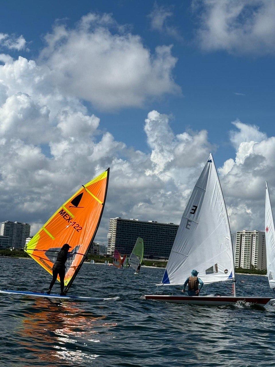 A colorful windsurfer and a small sailboat navigate the water under a partly cloudy sky with buildings in the background.$#$ CAPTION