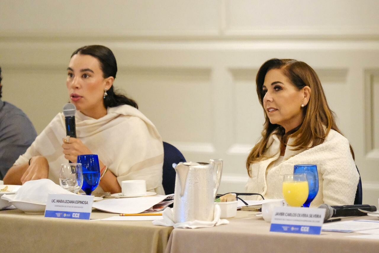 Two women seated at a conference table, one speaking into a microphone and the other listening attentively. Both are dressed in light-colored attire, with drinks and papers on the table.