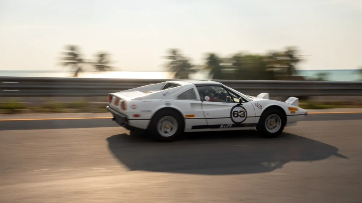 A white sports car with racing numbers speeds along a coastal road under a clear sky.$# CAPTION