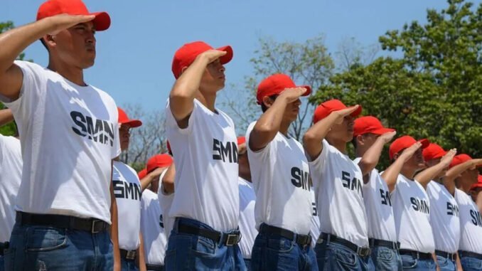 A group of young cadets in white t-shirts and red caps saluting in unison outdoors under a blue sky.