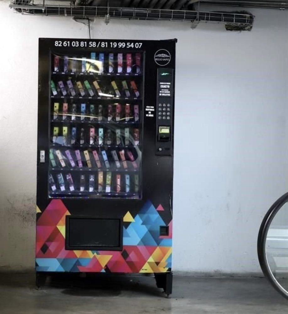 A vending machine filled with colorful products, located in an indoor area with a bicycle nearby. The machine has a geometric design and various selection buttons on the side.