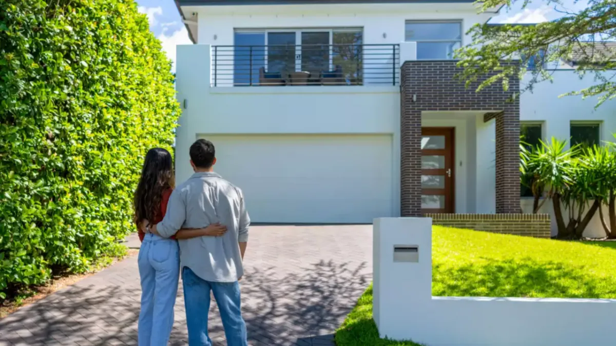 A couple stands together in front of a modern house, embracing and gazing at their new home. The house features a large garage and lush greenery surrounding it.