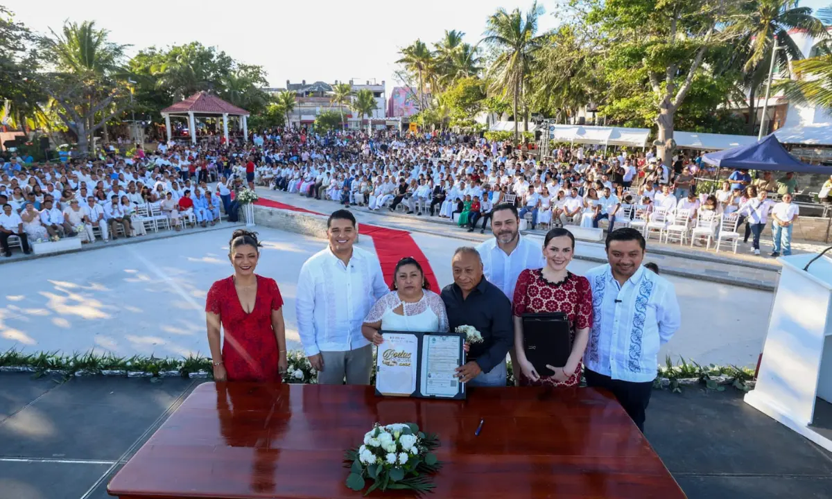 A large group of couples participating in a mass wedding ceremony at the Maya Portal in Playa del Carmen, with the sea visible in the background.