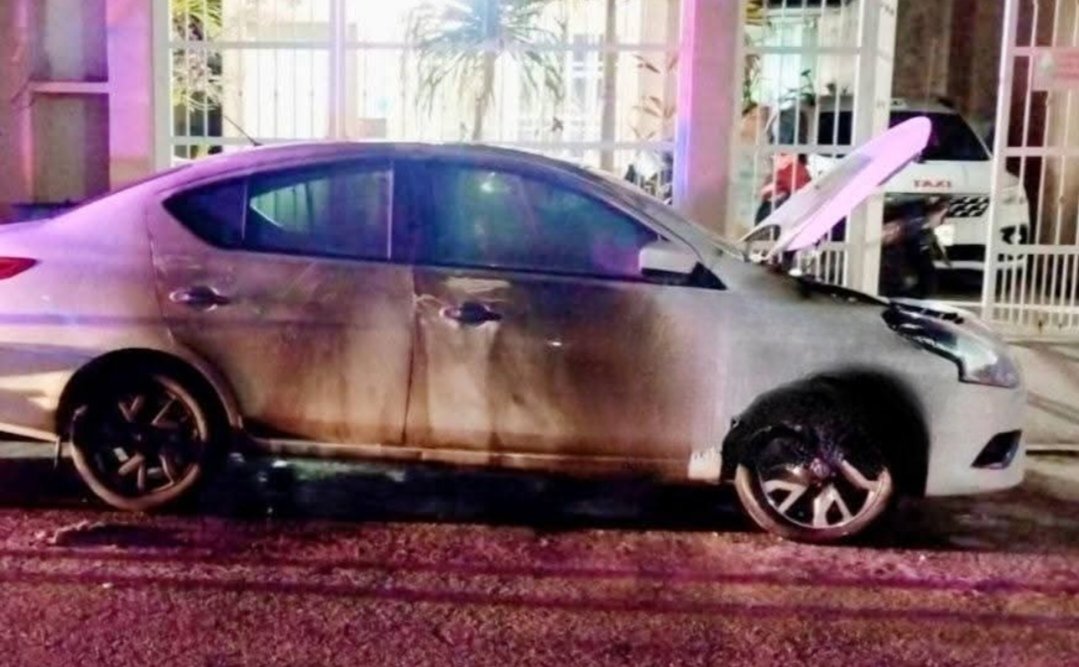 A silver car parked with its hood open, showing signs of neglect, in a nighttime setting with colorful lights in the background.