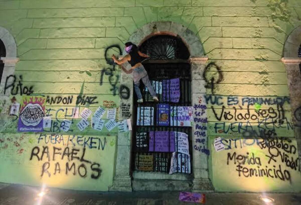 A person climbs on a wall covered with graffiti and posters related to social justice and feminism in a dimly lit urban environment.$# CAPTION