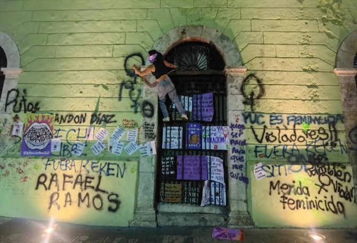 A person climbs on a wall covered with graffiti and posters related to social justice and feminism in a dimly lit urban environment.$# CAPTION