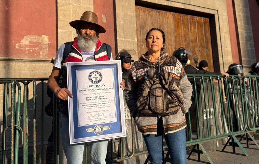 Two activists stand together holding a certificate from Guinness World Records in front of a closed building, with police visible in the background.$#$ CAPTION