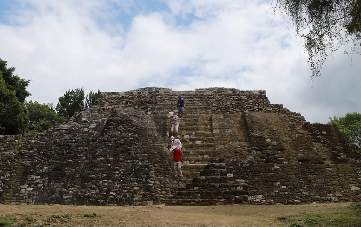 Visitors climbing the steps of a stone pyramid surrounded by greenery under a cloudy sky.$# CAPTION