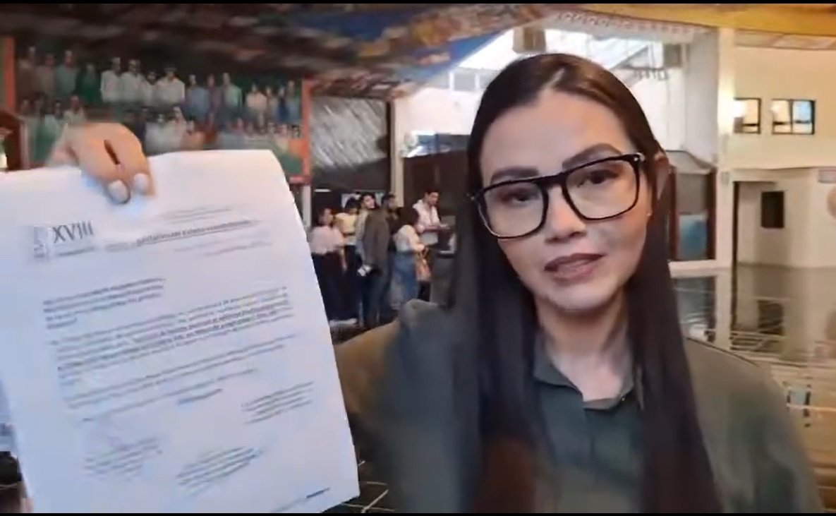 A woman holding a document in front of an audience in a hall, with a group of people in the background