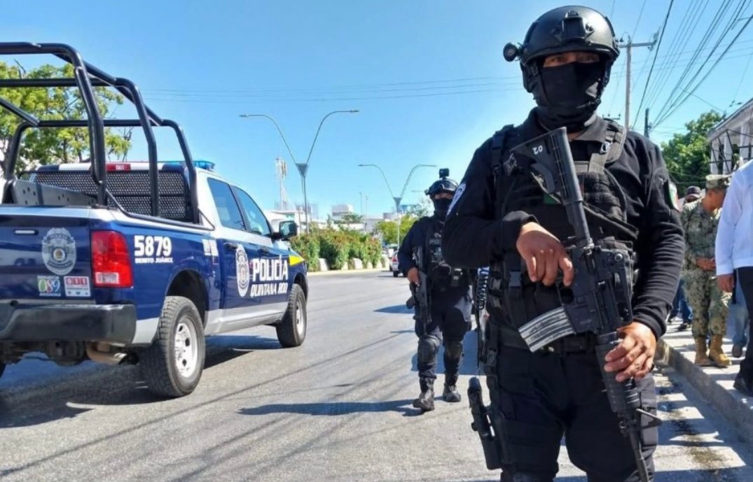 Heavily armed police officers patrol the streets of Quintana Roo, Mexico, with a police truck visible in the background.