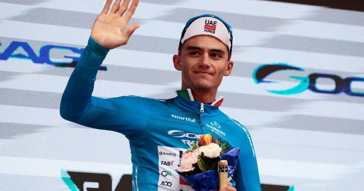 A cyclist in a blue jersey holds a bouquet while waving to the audience on a podium.$#$ CAPTION