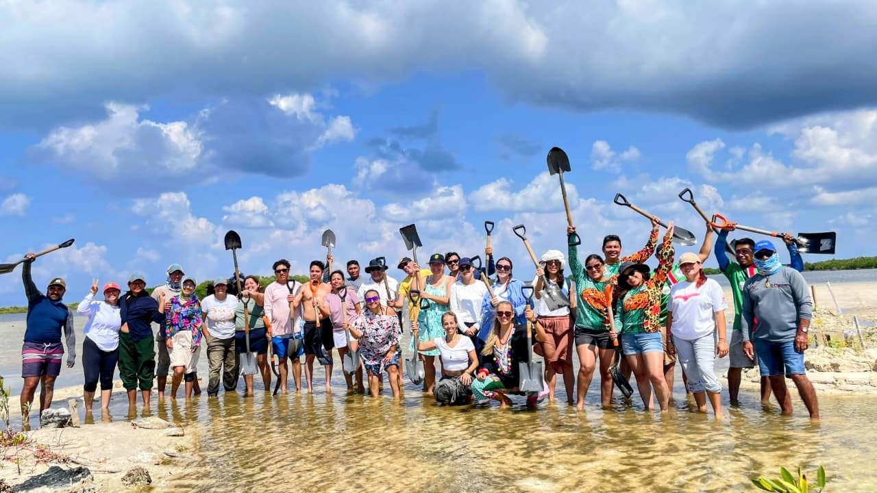 A large group of people, standing in shallow water, holding shovels and posing for a photo under a blue sky with clouds.