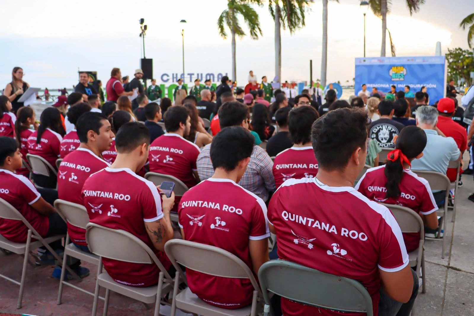 A crowd of people wearing matching shirts with "Quintana Roo" on the back, seated in chairs at an outdoor event, with palm trees in the background and a stage set up ahead of them.$#$ CAPTION