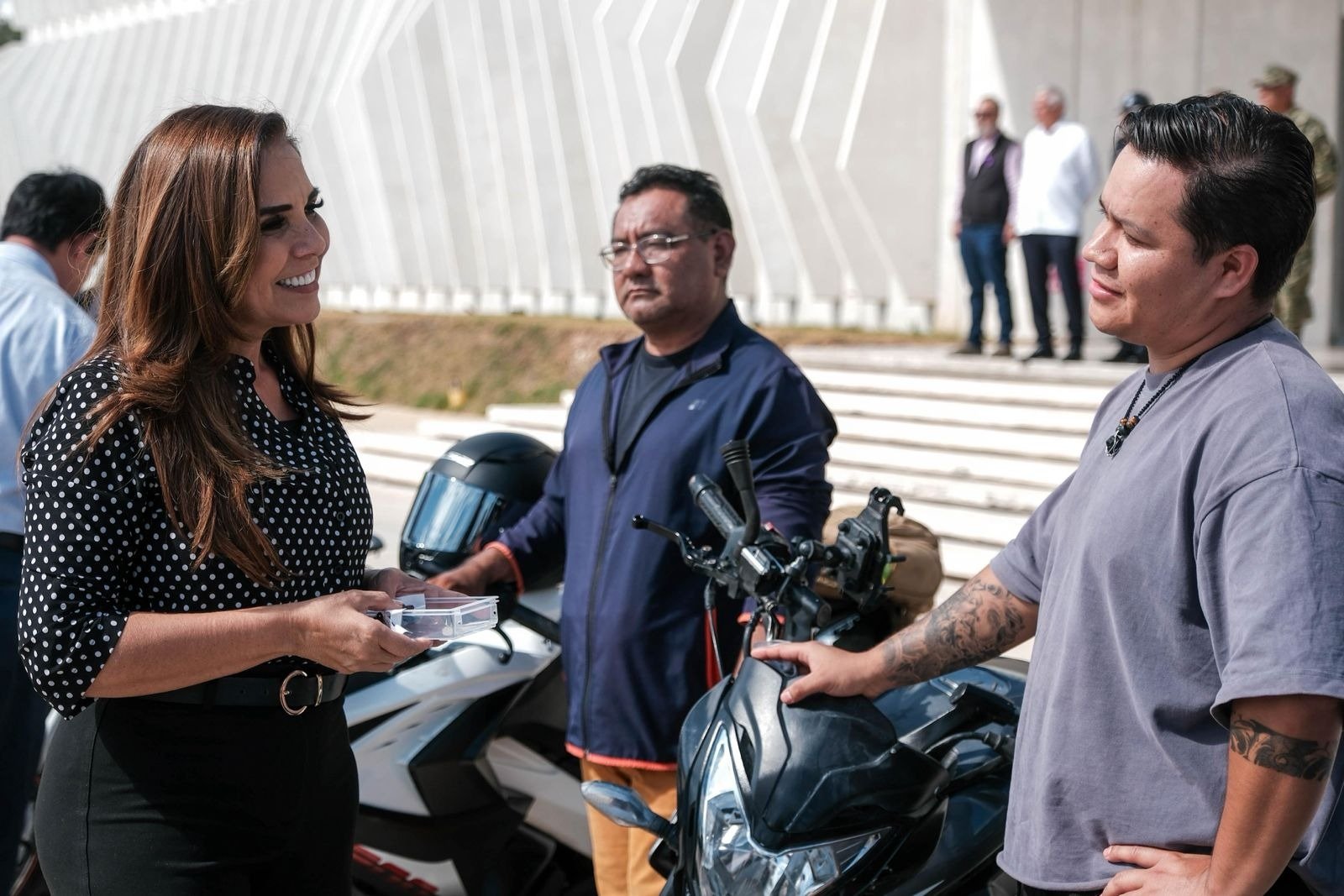 A group of three individuals engaged in conversation, with motorcycles in the background. One woman is holding a device, while two men listen attentively. The setting appears sunny with a modern architectural backdrop.$# CAPTION