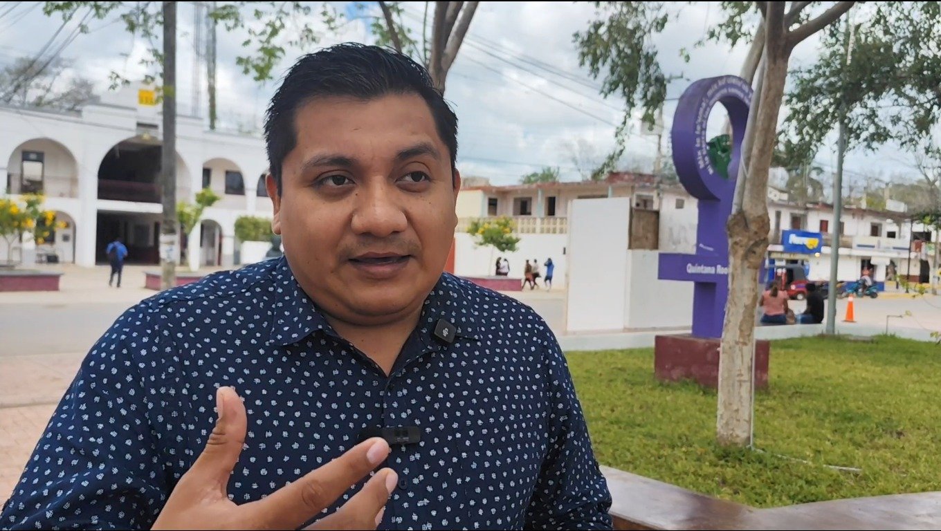 A man speaking while gesturing, with a purple symbol in the background, in a public area with people walking around - 31032026