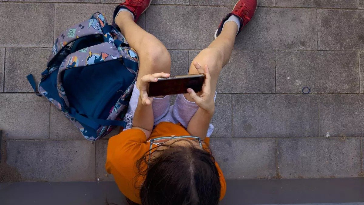 A young person sitting on the ground, holding a smartphone while wearing casual clothes and sneakers, with a backpack nearby.$# CAPTION