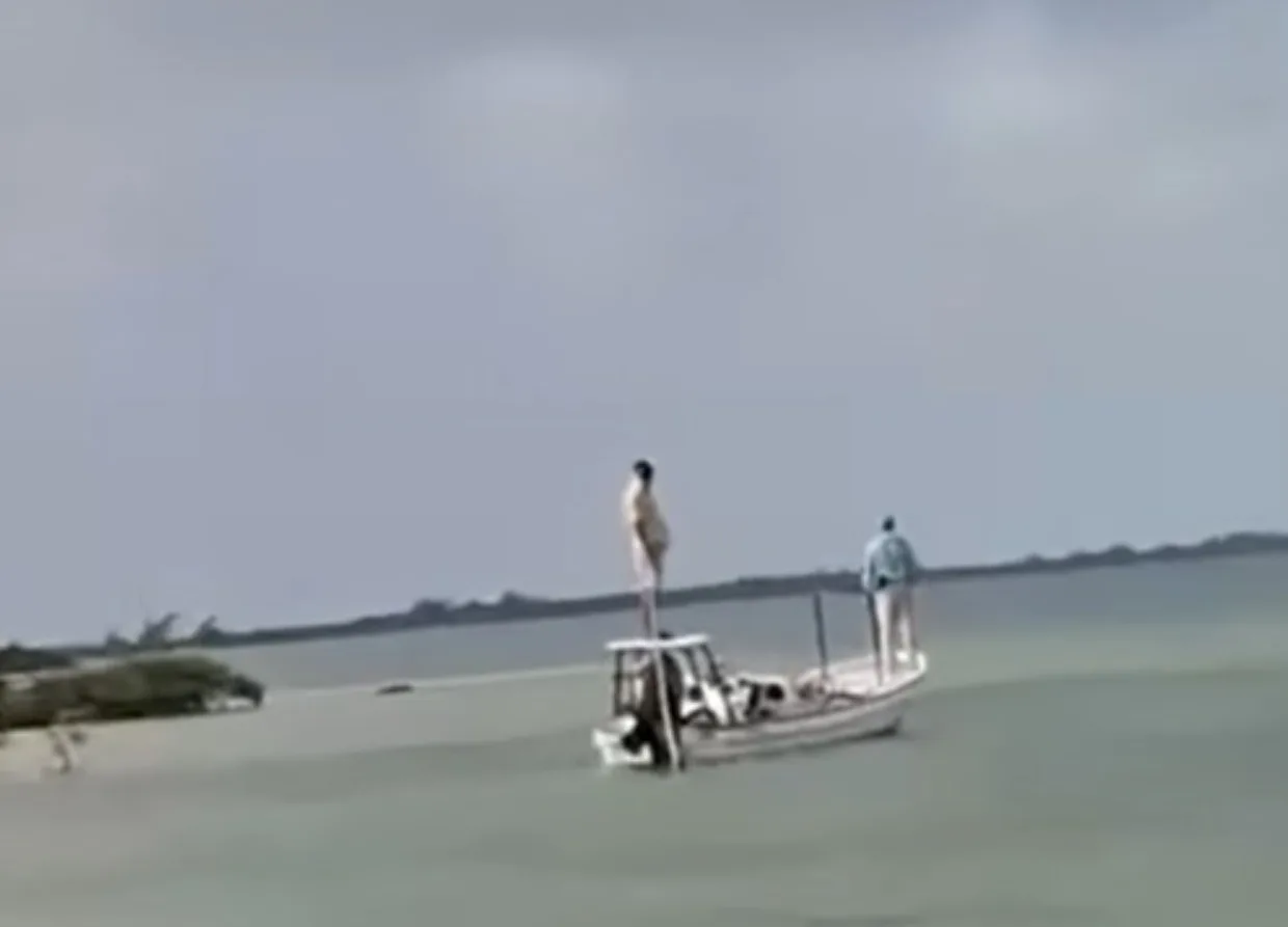 A small boat on calm water with two individuals, one standing on the bow and another at the stern, against a cloudy sky.$# CAPTION