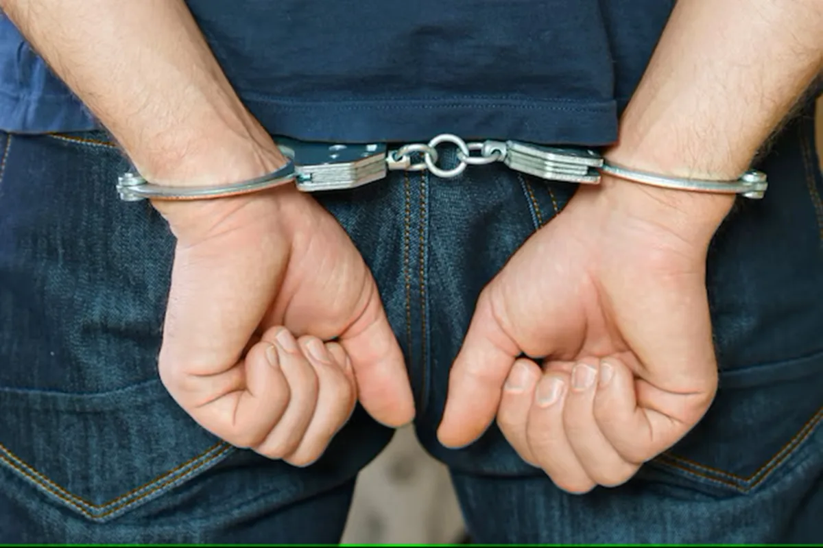 A person with their hands behind their back, wearing handcuffs. The individual is dressed in a blue shirt and denim jeans.