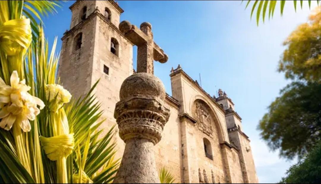 A historic church with stone walls, a cross, and blooming flowers in the foreground under a blue sky.