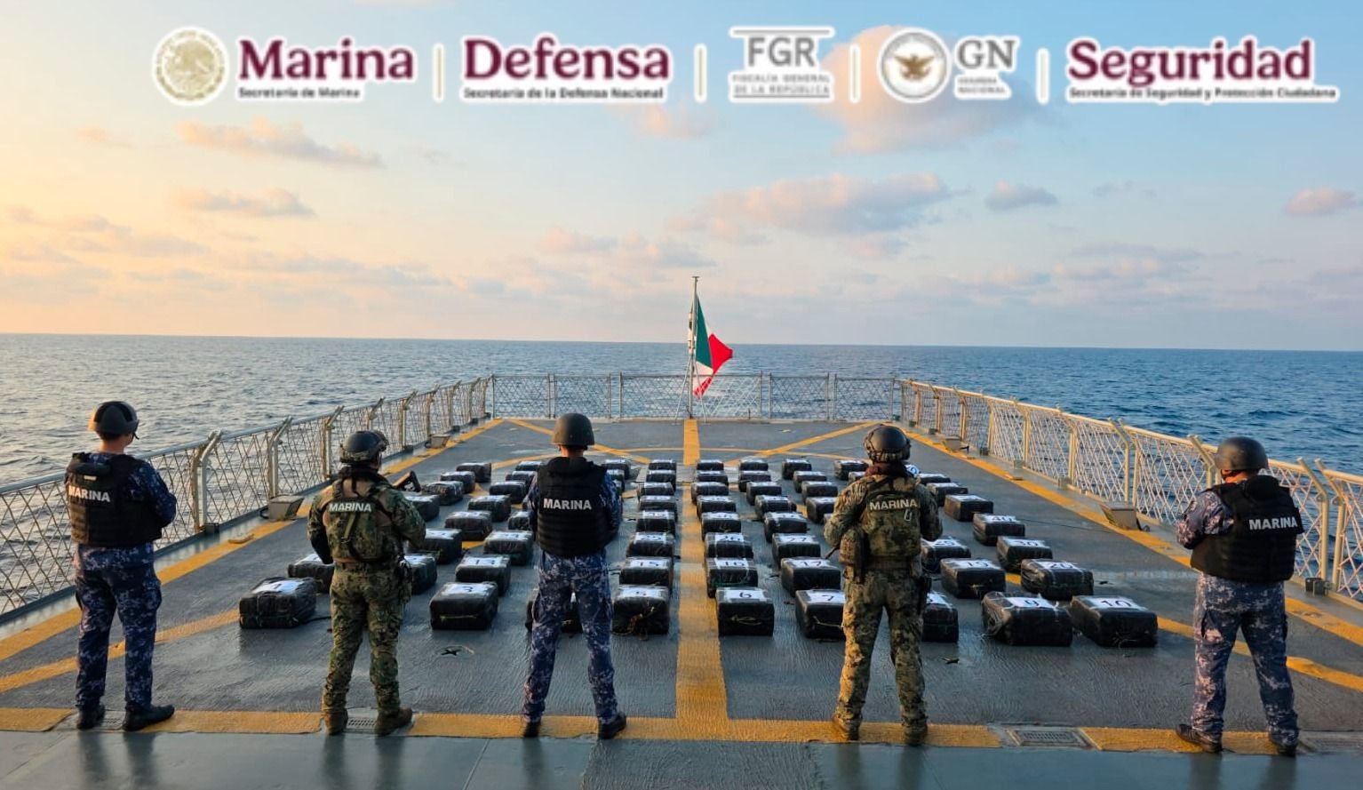 Mexican naval officers standing on a ship's deck, overlooking numerous packages laid out, with the ocean in the background and a Mexican flag in the center.