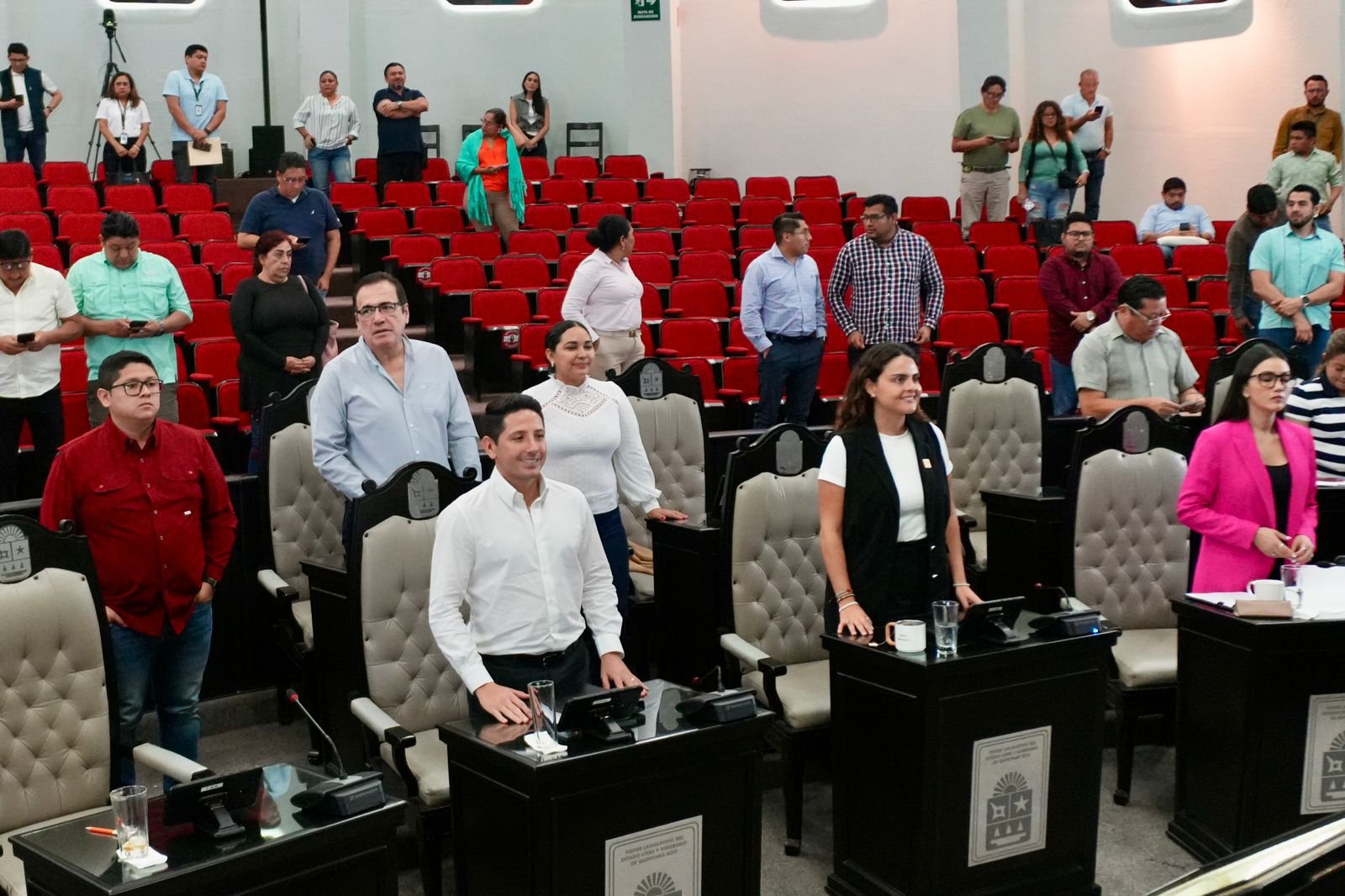 Members of a legislative assembly standing during a session, with red seats in the background and various attendees in the audience.$# CAPTION