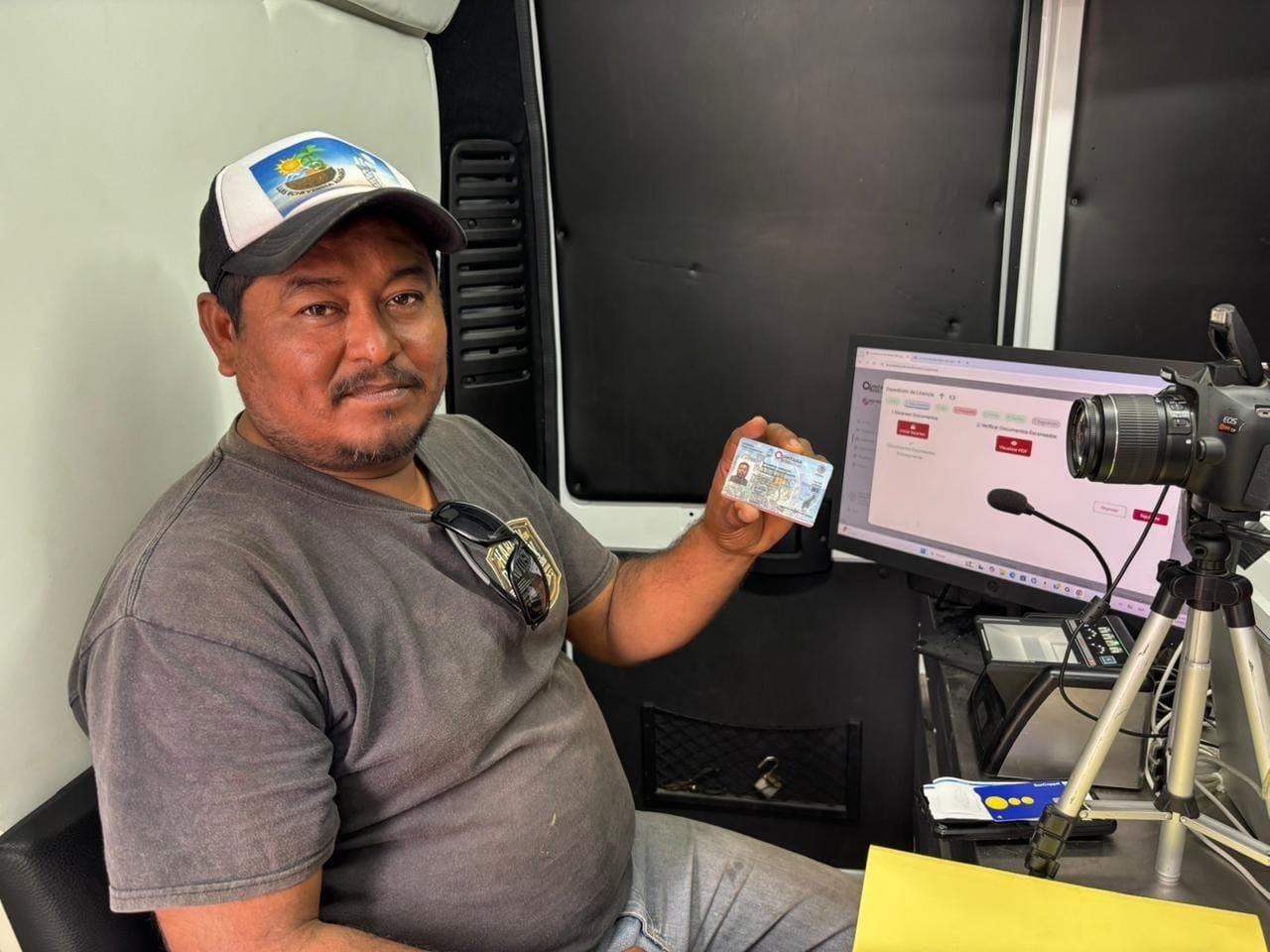 A man in a gray t-shirt and cap sits in a mobile office, holding an ID card, with a computer and camera in the background