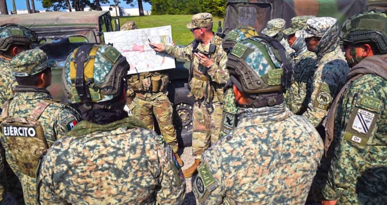 Soldiers in camouflage uniforms listen to a briefing while looking at a map in an outdoor setting.$# CAPTION