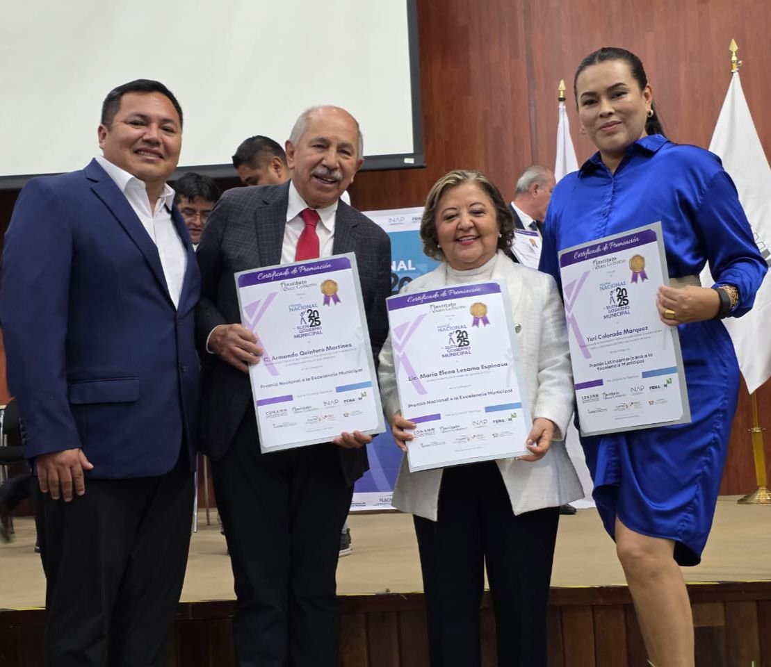 Four individuals holding certificates at an awards ceremony, with a backdrop of flags and banners in a formal setting