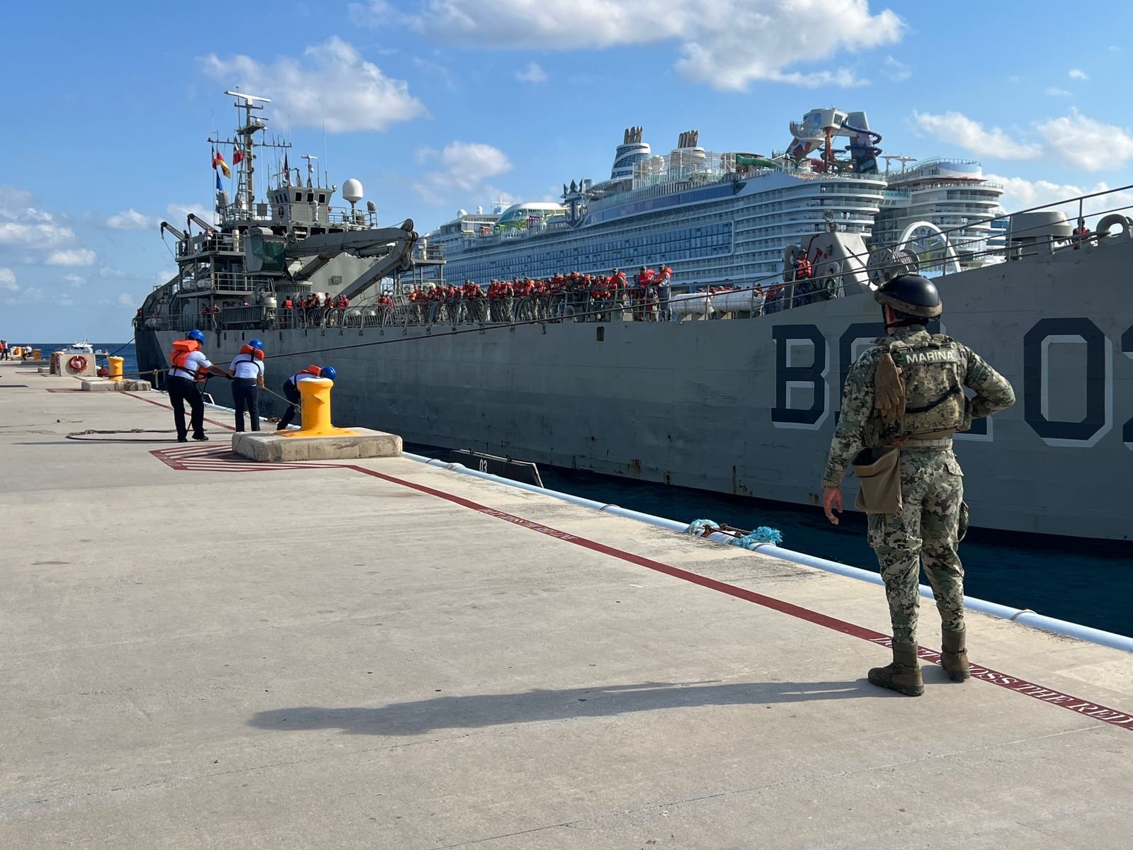 A naval ship docked at a port with crew members preparing to secure it. A soldier stands guard on the dock. In the background, a cruise ship is visible.