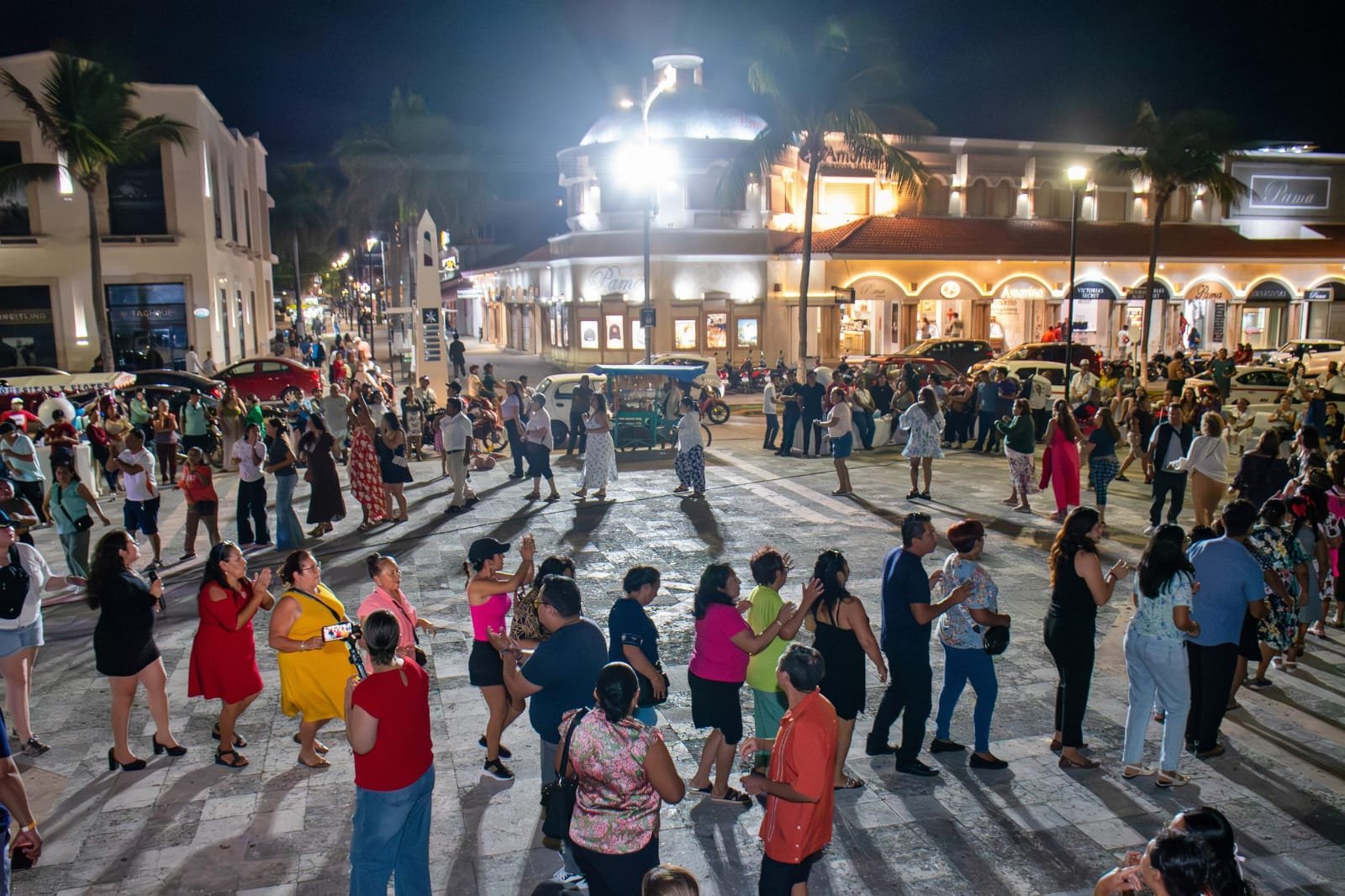 A vibrant outdoor scene of people dancing at night in a busy plaza, surrounded by palm trees and illuminated buildings.$# CAPTION