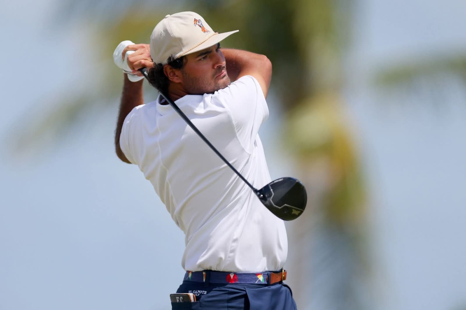 A golfer preparing to swing a club, focused and poised on the course with palm trees blurred in the background.